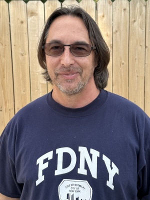 Jay Bingaman, Founder of Giocoso Studios: A headshot of Jay, a middle-aged man with medium-length brown hair, a short beard, and dark sunglasses. He is wearing a dark blue t-shirt with the white FDNY logo and is smiling slightly, with a wooden fence background.